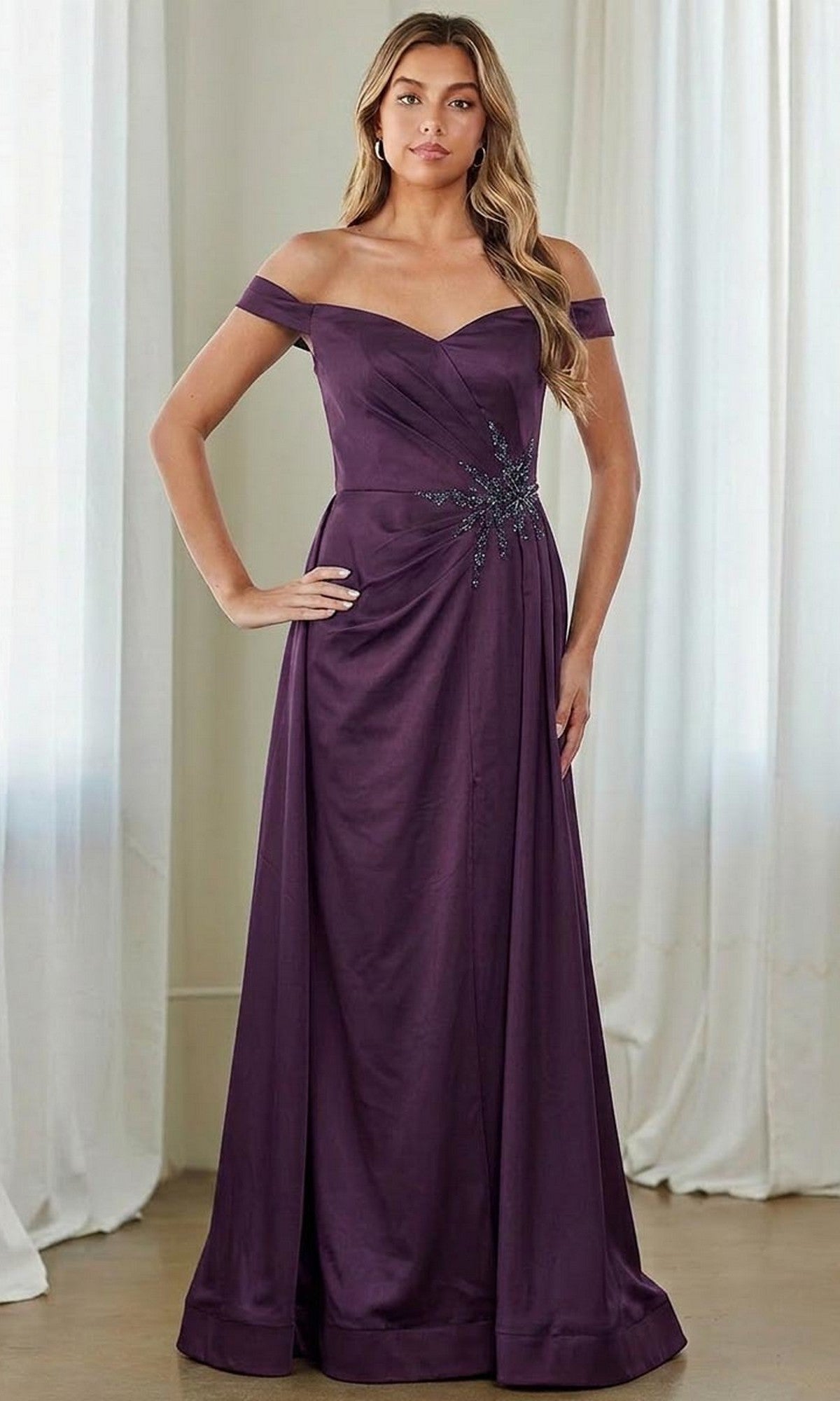 A woman stands indoors before white curtains, exuding timeless elegance in the Amelia Couture 7094—a floor-length eggplant off-the-shoulder formal dress with pleated details and a decorative waist, ideal as a mother of the bride gown.